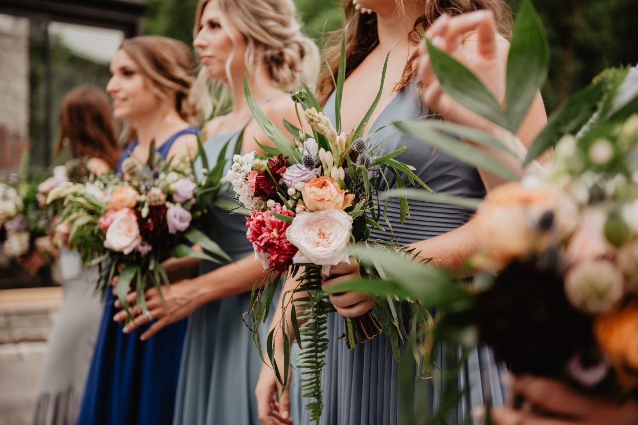 Focus on bouquet of flowers being held by bridesmaids in a line with them having similar dresses yet different, in shades of grey and darker blue