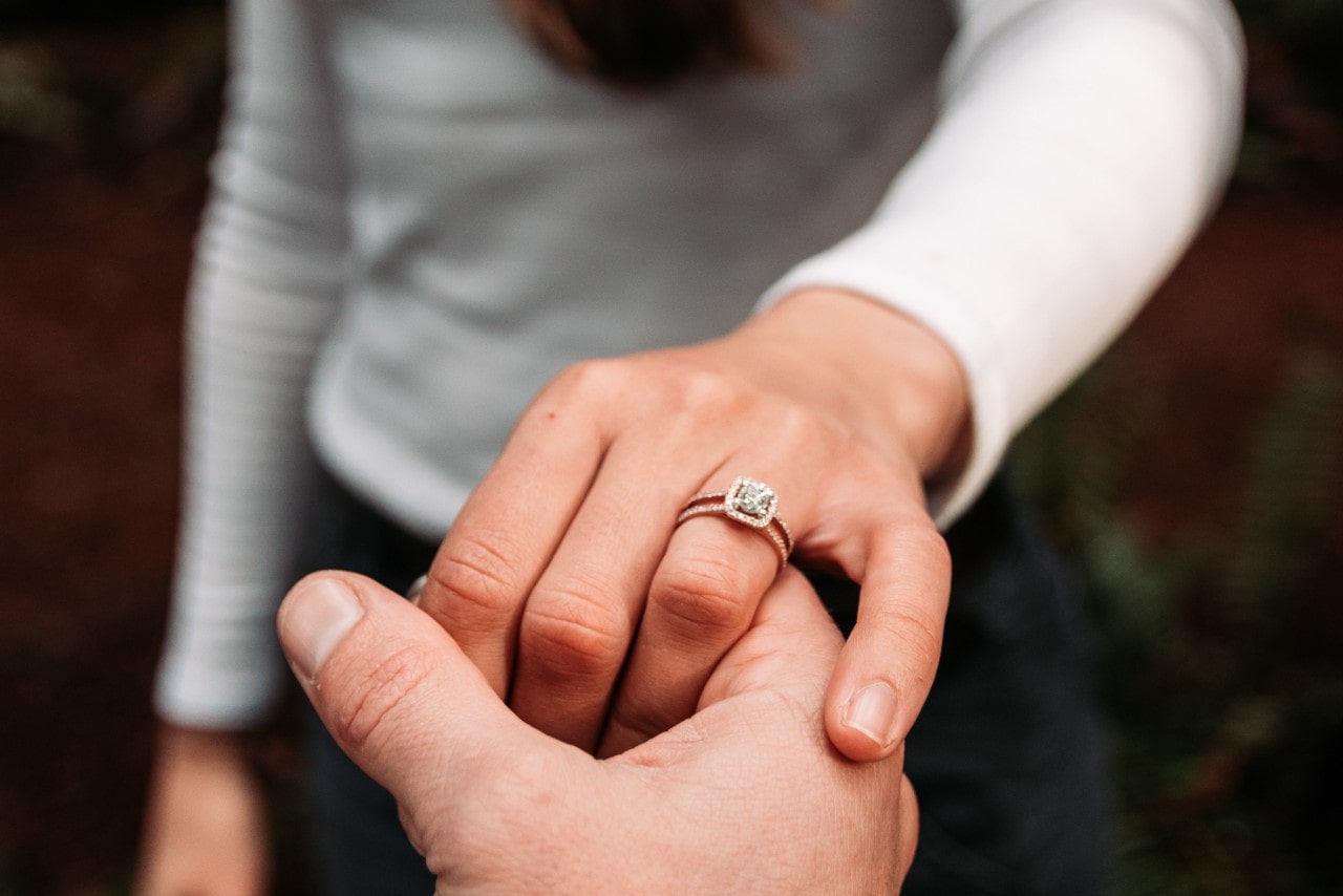 An engaged couple holding hands; the woman wearing a princess cut halo ring.