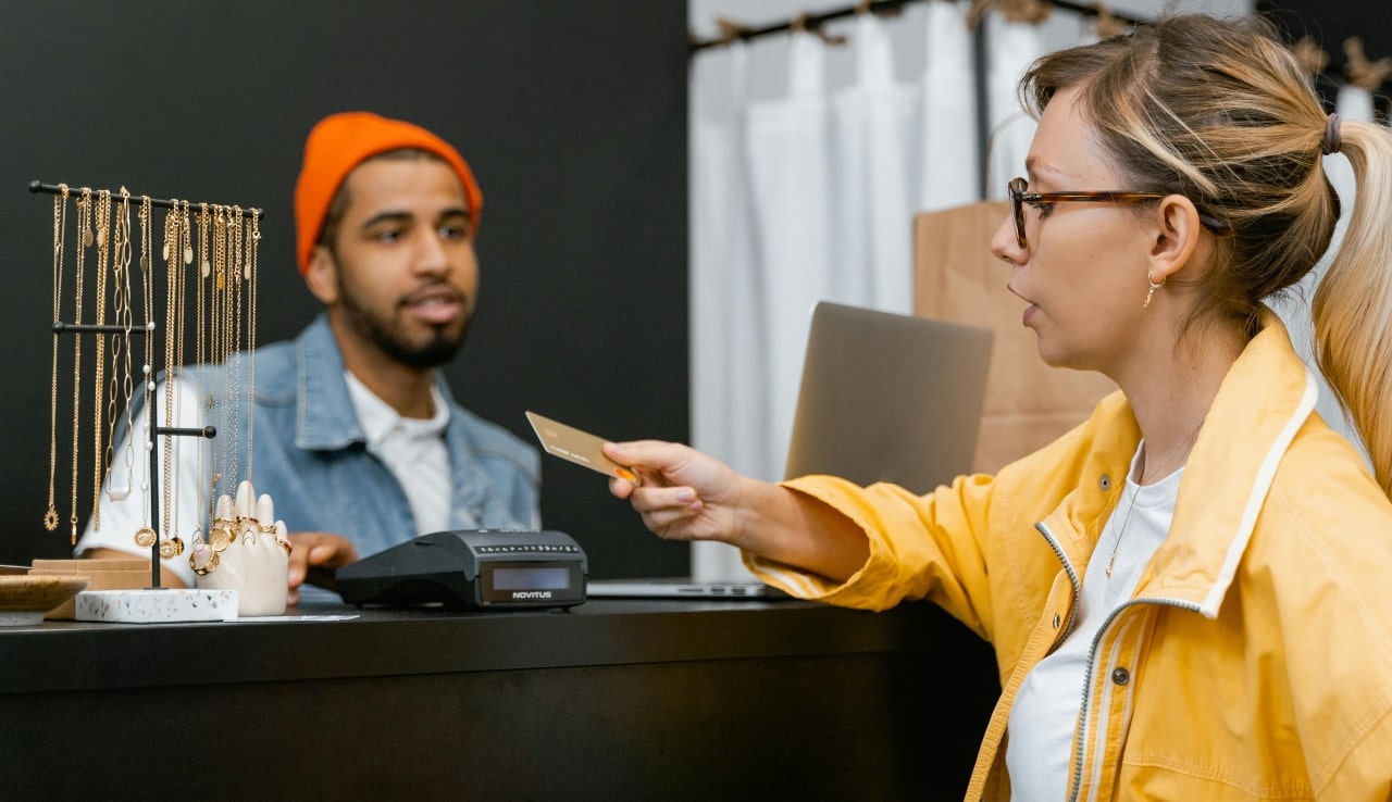 A woman in a yellow jacket is handing a credit card to a cashier at a jewelry store, with a display of gold necklaces and bracelets nearby.