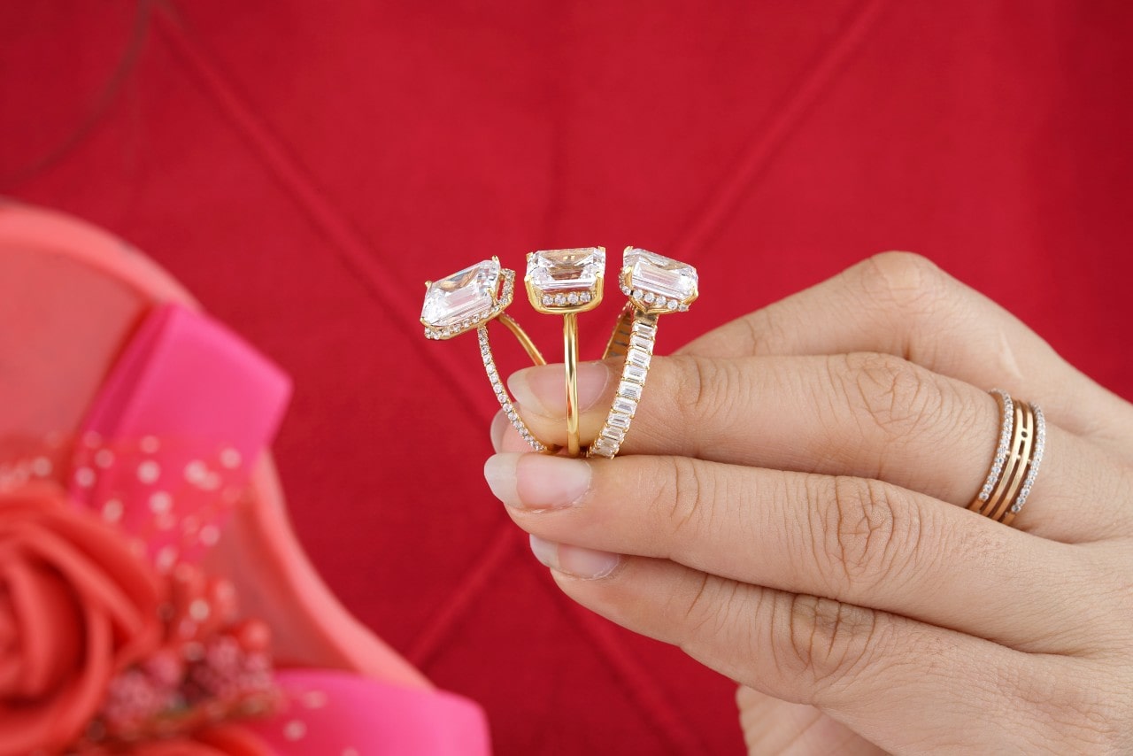 Closeup of a hand holding three diamond engagement rings against a red background