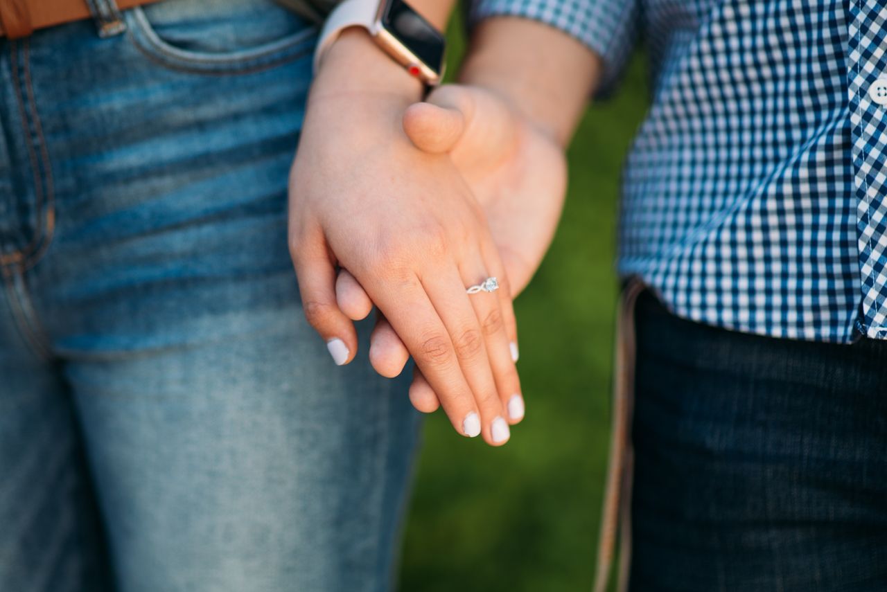 A man holds his fiance’s manicured hand to show off her diamond engagement ring after proposing on a casual nature hike